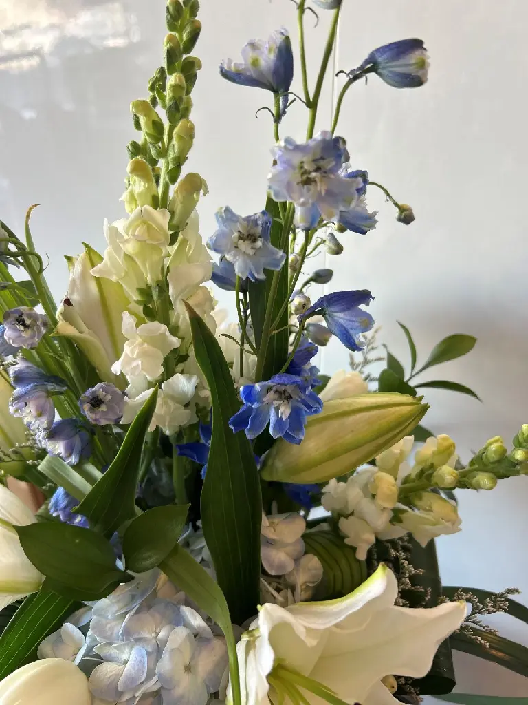 Composition florale blanche et bleue avec lys blancs, hortensias bleu pâle, delphiniums et feuillages, créée par La Maison d’Alcantara à Montréal.webp